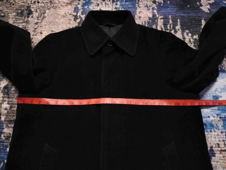 May include: A solid black coat with a pointed collar and long sleeves. A red measuring tape is placed horizontally across the chest, showing a width measurement of approximately 26 inches from one side to the other across the front of the garment.
