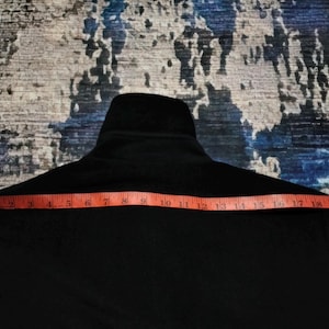 May include: A solid black jacket featuring a high collar and a long-sleeved design. An orange measuring tape is positioned horizontally across the chest area, indicating a width measurement of approximately 21 inches from edge to edge.