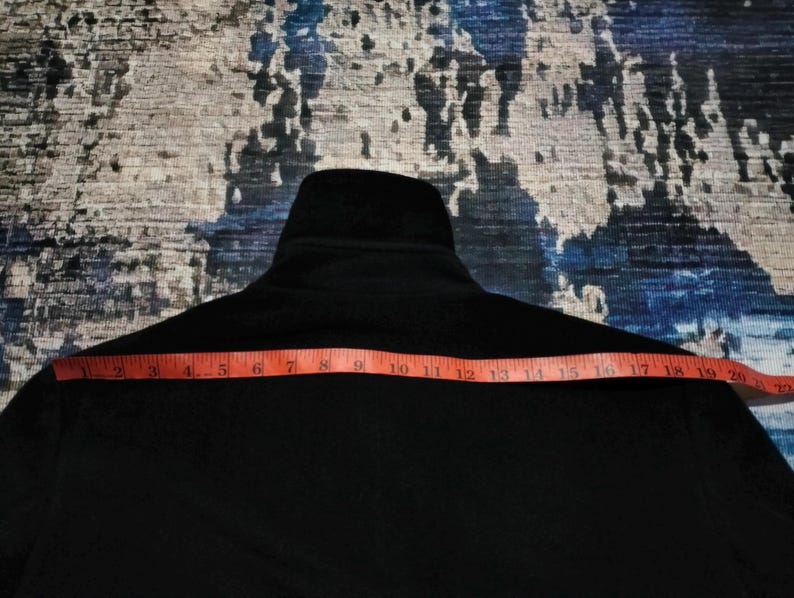 May include: A solid black jacket featuring a high collar and a long-sleeved design. An orange measuring tape is positioned horizontally across the chest area, indicating a width measurement of approximately 21 inches from edge to edge.