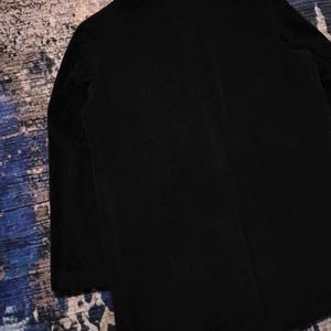 May include: A solid black blazer featuring a structured collar, long sleeves, and a clean, minimalist back design. The garment is crafted from a dark, matte fabric with visible vertical texture, showcasing a classic tailored silhouette and straight hem.