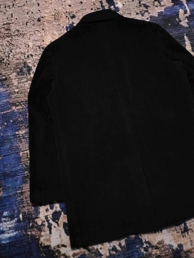 May include: A solid black blazer featuring a structured collar, long sleeves, and a clean, minimalist back design. The garment is crafted from a dark, matte fabric with visible vertical texture, showcasing a classic tailored silhouette and straight hem.