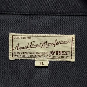 Puede incluir: Primer plano de una prenda azul marino con una etiqueta rectangular. La etiqueta dice "Armed Forces Manufacturer" con la marca "AVIREX" y el texto "AUTHENTIC GARMENT ESTABLISHED IN 1975". Tambi&eacute;n se ve la talla "3L".