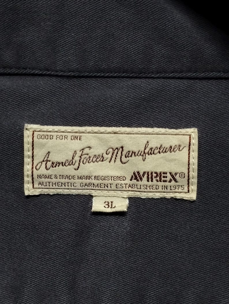 Puede incluir: Primer plano de una prenda azul marino con una etiqueta rectangular. La etiqueta dice "Armed Forces Manufacturer" con la marca "AVIREX" y el texto "AUTHENTIC GARMENT ESTABLISHED IN 1975". Tambi&eacute;n se ve la talla "3L".