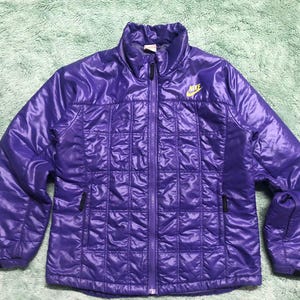 May include: A purple quilted jacket with a zip fastening and a Nike logo on the right chest. The jacket has a shiny finish.