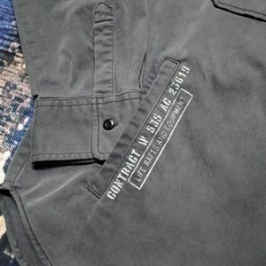 Puede incluir: Una camisa gris oscuro con botones, un bolsillo y mangas largas. El bolsillo tiene texto blanco que dice "CONTRACT W 535 AC 25619 LIFE RAFTS AND EQUIPMENT". La camisa tiene un solo bot&oacute;n negro en el pu&ntilde;o.
