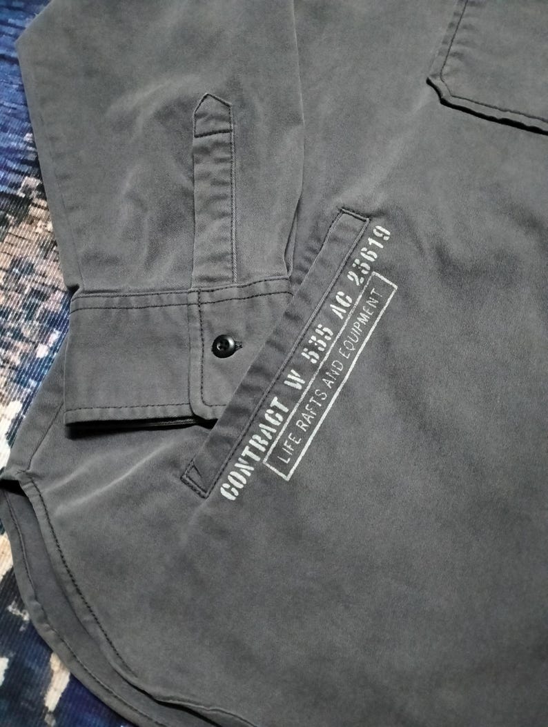 Puede incluir: Una camisa gris oscuro con botones, un bolsillo y mangas largas. El bolsillo tiene texto blanco que dice "CONTRACT W 535 AC 25619 LIFE RAFTS AND EQUIPMENT". La camisa tiene un solo bot&oacute;n negro en el pu&ntilde;o.