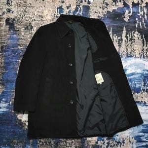 May include: A black coat with a classic collar and a front button closure. The interior is lined with a dark fabric and features a white tag with the text A.A.R printed in black. The coat has long sleeves and a structured, straight-cut silhouette.