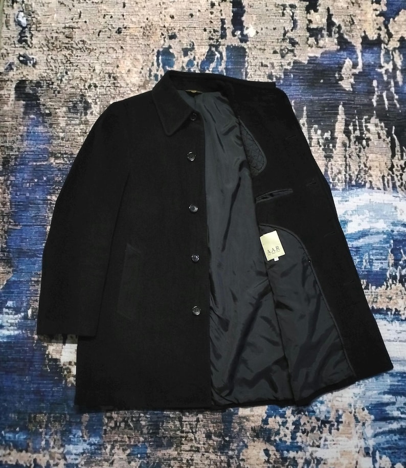 May include: A black coat with a classic collar and a front button closure. The interior is lined with a dark fabric and features a white tag with the text A.A.R printed in black. The coat has long sleeves and a structured, straight-cut silhouette.