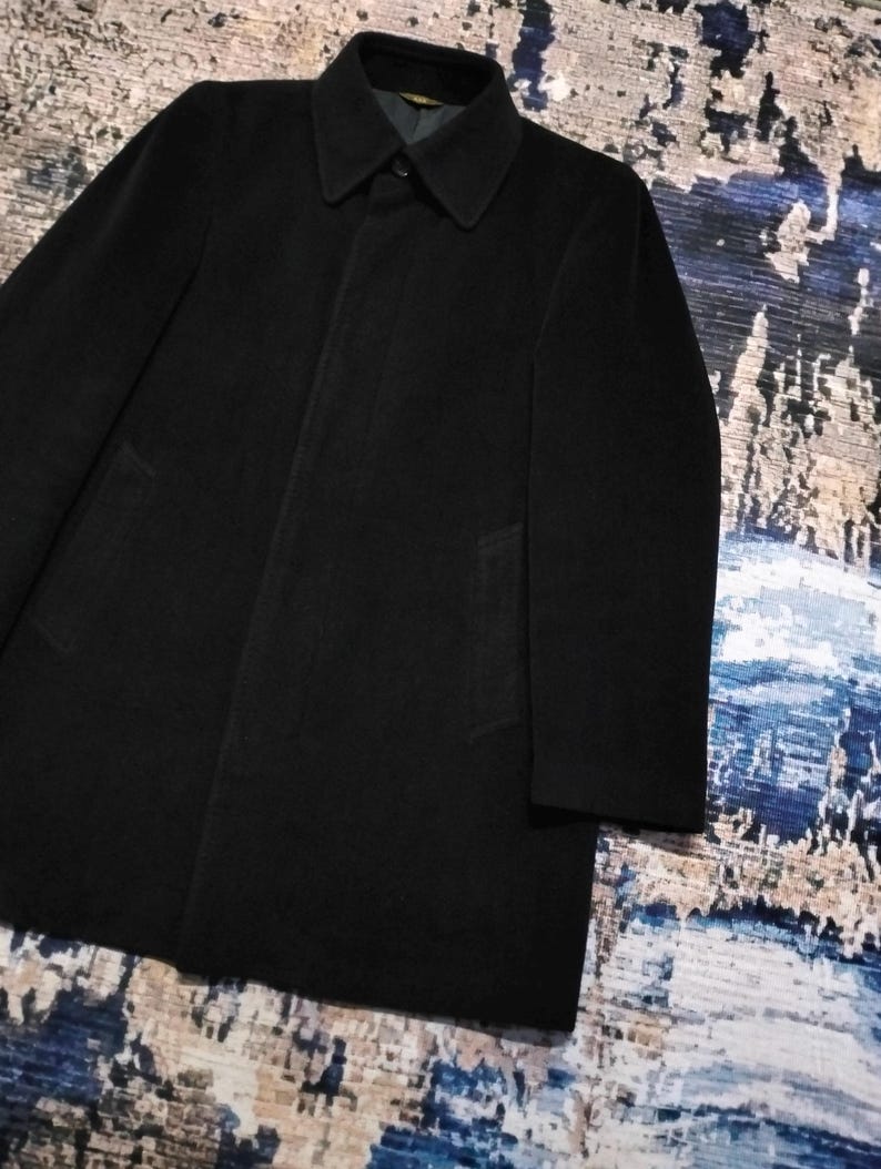 May include: A solid black overcoat featuring a structured collar, long sleeves, and a concealed button placket. The coat has a smooth, matte finish and a clean, straight silhouette that extends to the mid-thigh, providing a classic and simple look.