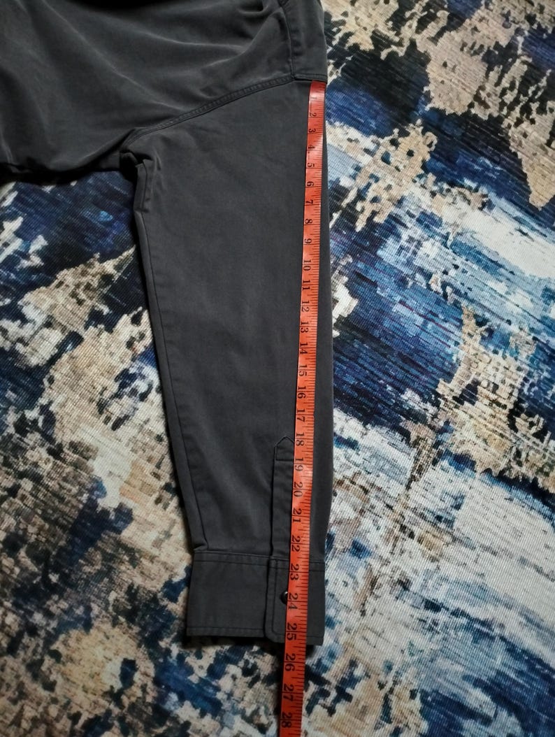 Puede incluir: Una camisa de manga larga gris oscuro con pu&ntilde;o abotonado. Una cinta m&eacute;trica roja y blanca se coloca a lo largo de la manga, mostrando medidas de hasta 71 cm.