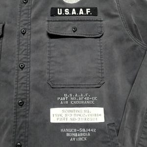 Puede incluir: Camisa gris oscuro con botones, bolsillo en el pecho y el texto "U.S.A.A.F." encima. La camisa presenta un emblema de ala blanca y texto adicional, incluyendo "AIR ENDURANCE" y "AVIREX".