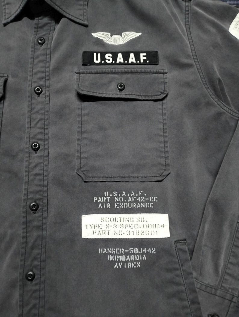 Puede incluir: Camisa gris oscuro con botones, bolsillo en el pecho y el texto "U.S.A.A.F." encima. La camisa presenta un emblema de ala blanca y texto adicional, incluyendo "AIR ENDURANCE" y "AVIREX".