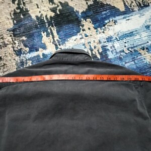 Puede incluir: Una camisa gris oscuro con botones y cuello, medida con una cinta m&eacute;trica roja. La cinta m&eacute;trica muestra una medida de aproximadamente 56 cm en los hombros. La camisa est&aacute; sobre una superficie azul y gris estampada.