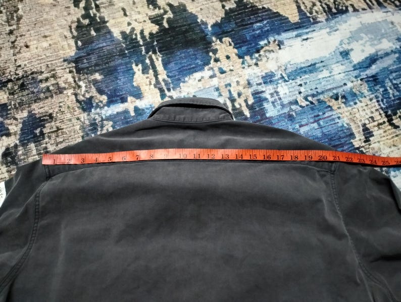 Puede incluir: Una camisa gris oscuro con botones y cuello, medida con una cinta m&eacute;trica roja. La cinta m&eacute;trica muestra una medida de aproximadamente 56 cm en los hombros. La camisa est&aacute; sobre una superficie azul y gris estampada.