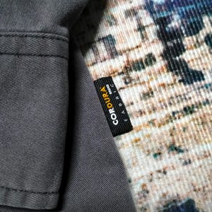 Puede incluir: Primer plano de una tela gris con una etiqueta negra que dice "CORDURA FABRIC BRAND" en texto blanco. La tela parece ser parte de una prenda, con costuras visibles. El fondo presenta un textil estampado.