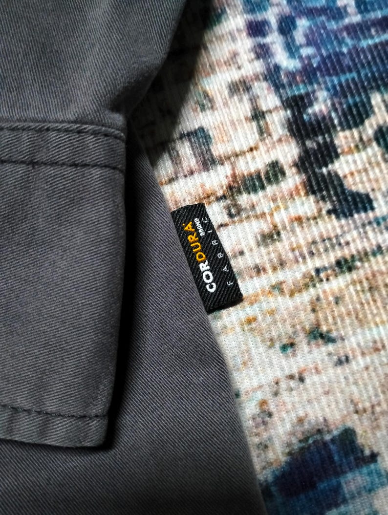 Puede incluir: Primer plano de una tela gris con una etiqueta negra que dice "CORDURA FABRIC BRAND" en texto blanco. La tela parece ser parte de una prenda, con costuras visibles. El fondo presenta un textil estampado.