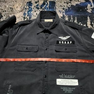 Puede incluir: Camisa gris oscuro con botones, dos bolsillos en el pecho y charreteras. Cuenta con un parche de U.S.A.A.F. y texto en la parte inferior delantera. Una cinta m&eacute;trica roja est&aacute; colocada sobre la camisa. La camisa tiene mangas largas y cuello.