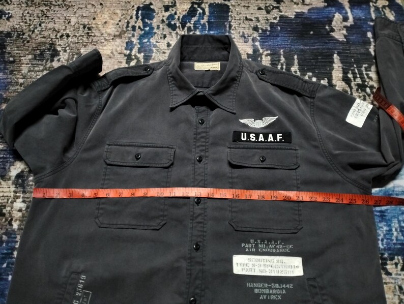 Puede incluir: Camisa gris oscuro con botones, dos bolsillos en el pecho y charreteras. Cuenta con un parche de U.S.A.A.F. y texto en la parte inferior delantera. Una cinta m&eacute;trica roja est&aacute; colocada sobre la camisa. La camisa tiene mangas largas y cuello.