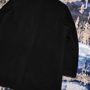 May include: A solid black coat featuring a structured collar, long sleeves, and a single vertical seam running down the center of the back. The garment is crafted from a dark, matte fabric with a smooth texture, showcasing a clean and simple design.