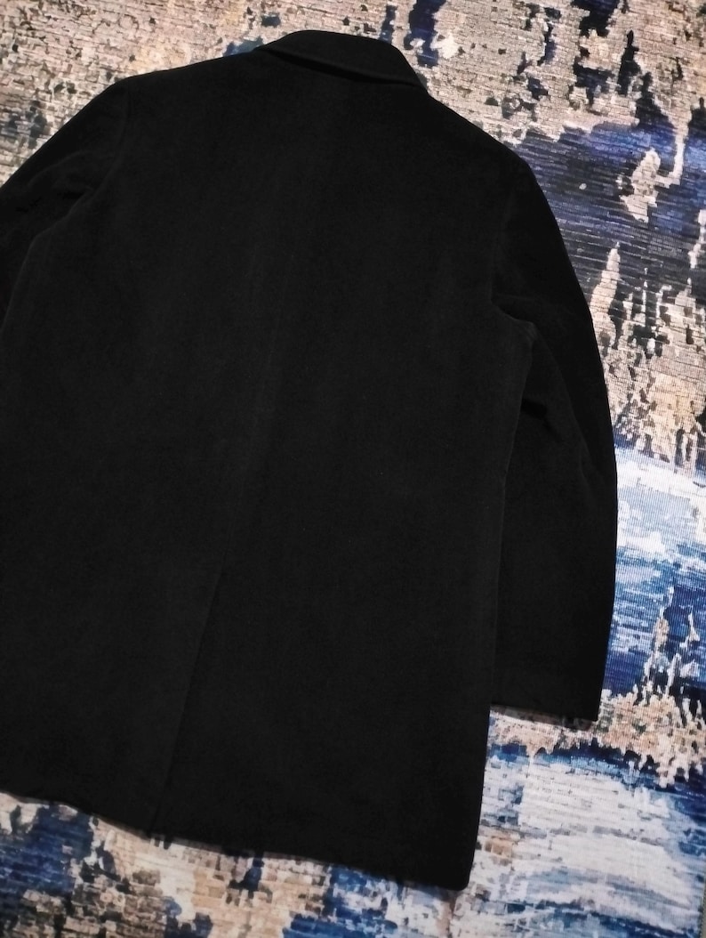 May include: A solid black coat featuring a structured collar, long sleeves, and a single vertical seam running down the center of the back. The garment is crafted from a dark, matte fabric with a smooth texture, showcasing a clean and simple design.
