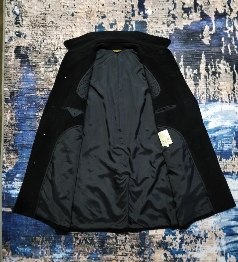 May include: A black coat with a wide collar and long sleeves, shown open to reveal a dark interior lining. The garment features quilted shoulder panels and a small white rectangular tag on the inside right panel with the text AAR printed on it.