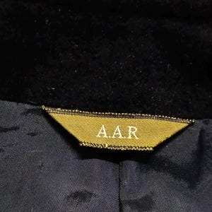 May include: A black garment featuring a triangular gold-colored fabric label sewn into the neckline. The label displays the text A.A.R in a light-colored font. The garment has a dark blue interior lining with a visible seam running vertically.