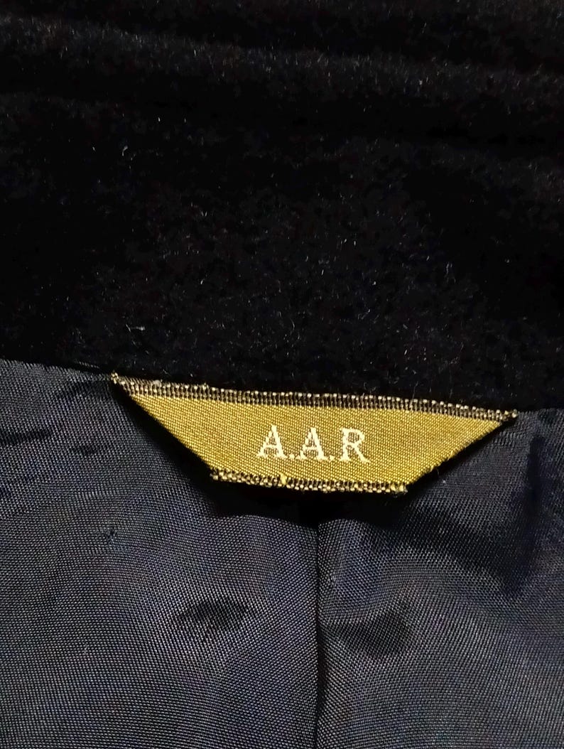 May include: A black garment featuring a triangular gold-colored fabric label sewn into the neckline. The label displays the text A.A.R in a light-colored font. The garment has a dark blue interior lining with a visible seam running vertically.