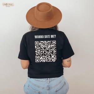May include: Black t-shirt with the text "Wanna Date Me?" and a black and white QR code printed on the back. The shirt is being worn by a person wearing a brown hat and blue jeans.