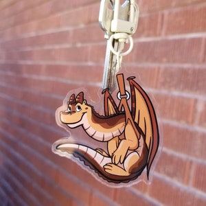 Puede incluir: Un llavero de dragón de dibujos animados marrón y naranja con una cara sonriente. El dragón está sujetando una correa marrón con un anillo plateado.