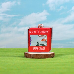May include: A red acrylic keychain shaped like a fire alarm box with the text "In case of sadness, break glass". The box has a clear window with a cartoon image of a cat and a dog inside.