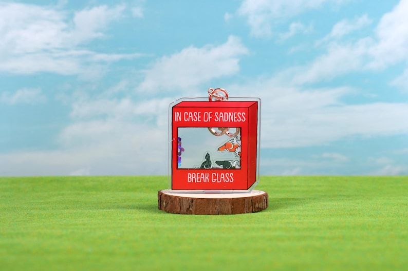 May include: A red acrylic keychain shaped like a fire alarm box with the text "In case of sadness, break glass". The box has a clear window with a cartoon image of a cat and a dog inside.