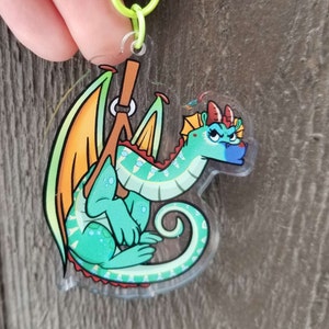 May include: A clear acrylic charm featuring a cartoon dragon with green scales, red and orange wings, and a blue and white face. The dragon is holding a brown stick with a ring on it.