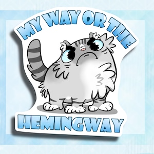 以下が含まれることがあります： 不機嫌そうな表情をした灰色と白の猫が上を見ている。猫は「My Way Or The Hemingway」という文字が書かれた青と白のステッカーに囲まれている。