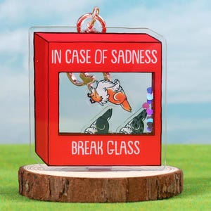May include: A red acrylic keychain shaped like a fire alarm with the text "In case of sadness break glass". Inside the glass is a cartoon image of a dog with a red and white patterned body and two other dogs with black and white patterned bodies.