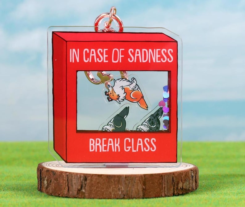 May include: A red acrylic keychain shaped like a fire alarm with the text "In case of sadness break glass". Inside the glass is a cartoon image of a dog with a red and white patterned body and two other dogs with black and white patterned bodies.