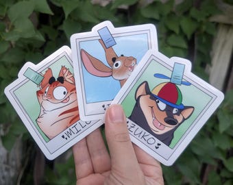 May include: Three square stickers designed like polaroid photos. One features an orange tabby cat labeled MILO, one shows a brown rabbit, and one depicts a black and tan dog wearing a propeller hat labeled ZUKO. Each has a small blue tab at the top.