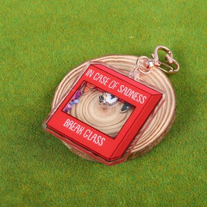 May include: Red acrylic keychain with the text "In case of sadness, break glass". The keychain features a small, colorful scene inside the glass.