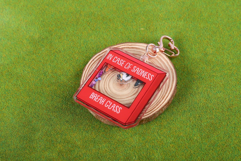 May include: Red acrylic keychain with the text "In case of sadness, break glass". The keychain features a small, colorful scene inside the glass.