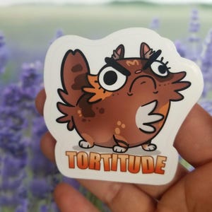 May include: A brown and orange cartoon cat with a grumpy expression and the word "Tortitude" written below it. The cat is wearing a white sticker.