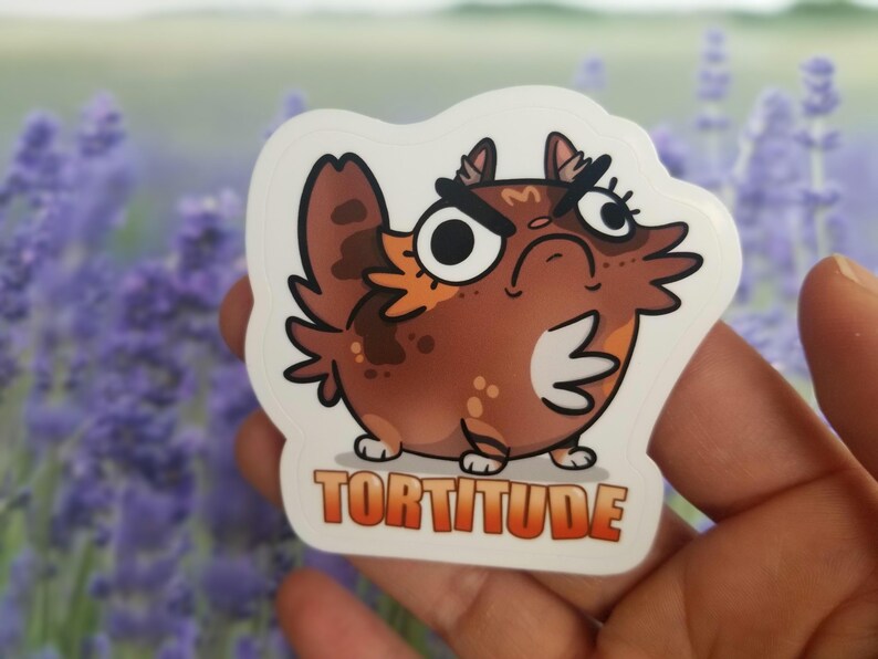 May include: A brown and orange cartoon cat with a grumpy expression and the word "Tortitude" written below it. The cat is wearing a white sticker.