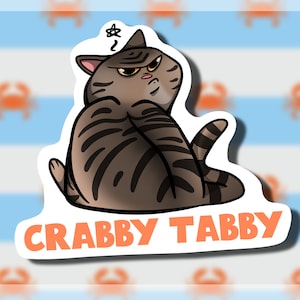 May include: A sticker featuring a cartoon tabby cat with a grumpy expression. The cat is gray and black with stripes. The sticker has the text "Crabby Tabby" in orange letters.