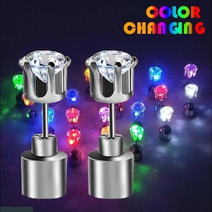 May include: Two silver-colored stud earrings with clear, faceted tops. The earrings have a cylindrical base and a post. The background features multiple color-changing LED lights. The text "COLOR CHANGING" is displayed in rainbow colors.