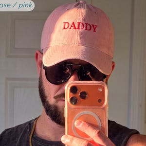 May include: A dusty rose pink baseball cap featuring the word DADDY embroidered in red thread on the front panel. The cap has a structured crown and a curved brim. A small label in the top left corner displays the text dusty rose / pink.