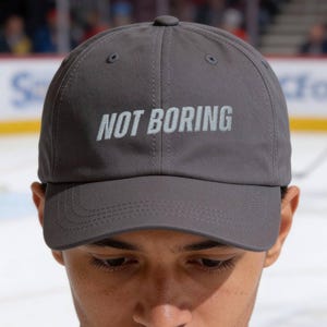 May include: A gray baseball cap with the text "NOT BORING" in white block letters. The cap is made of a soft material and has a curved brim. The cap is being worn by a person.