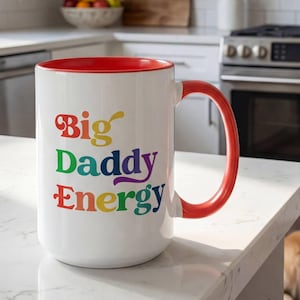 May include: A white ceramic mug with a bright red interior and handle. The exterior features the text "Big Daddy Energy" printed in a multi-colored, retro-style font with letters in red, yellow, green, purple, and blue, positioned on a kitchen counter.