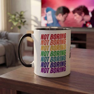May include: A white ceramic mug with a black handle and interior. The exterior features the text NOT BORING repeated seven times in a vertical stack, with each line printed in a different color of the rainbow: red, orange, yellow, green, blue, indigo, and violet.