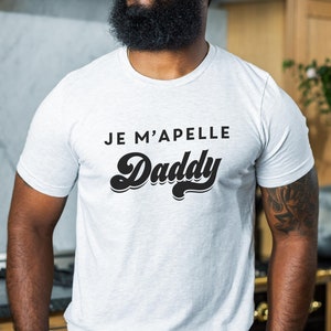 Puede incluir: Camiseta gris claro con el texto "JE M'APELLE Daddy" en negro. La palabra "Daddy" está en una fuente retro y audaz. La camiseta es de manga corta y está hecha de un material suave.