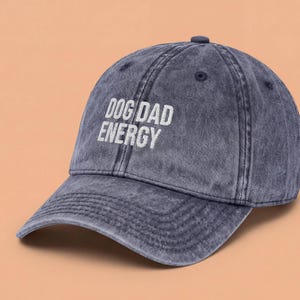 May include: A navy blue baseball cap with a vintage-style acid-washed finish. The front of the cap features the embroidered white text DOG DAD ENERGY in a clean, sans-serif font. The hat has a curved brim and a structured six-panel crown design.