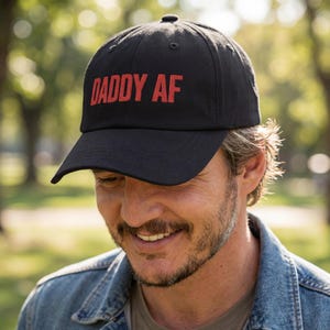 May include: A black baseball cap featuring the text DADDY AF embroidered in red capital letters across the front panel. The cap has a structured crown, a curved brim, and a button at the top center, providing a classic look with bold lettering.