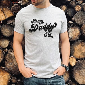 May include: A grey t-shirt with the text "In my Daddy Era" in a retro font.